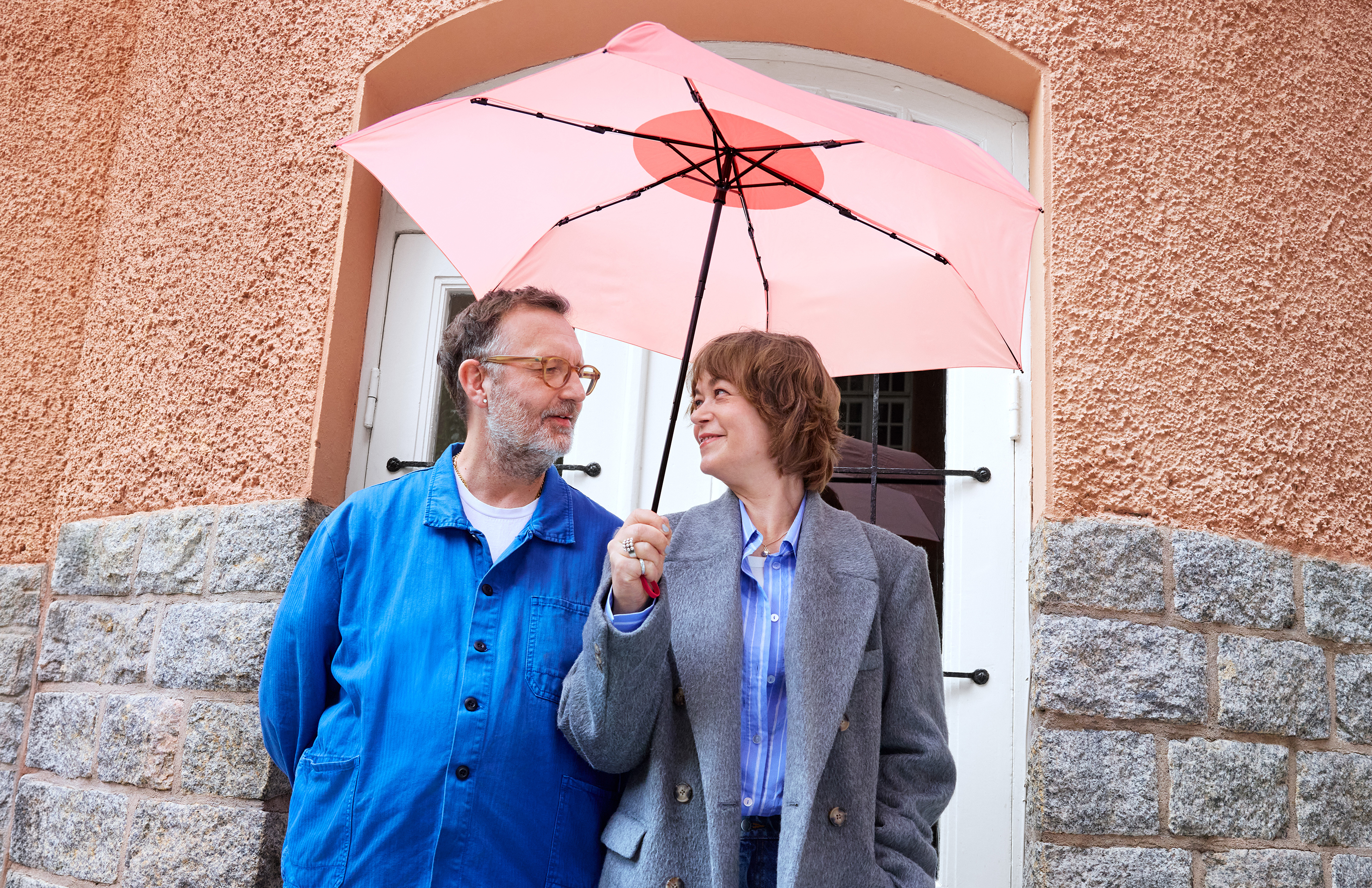 Två personer under Lindex rosa paraply Pink Umbrella framför en orange fasad